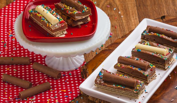 Foto de uma bancada de madeira, sobre ela há um pano vermelho com bolinhas brancas, um prato retangular com a receita pronta, alguns chocolates Baton soltos e mais um prato vermelho elevado. Há alguns granulados coloridos decorando a cena.