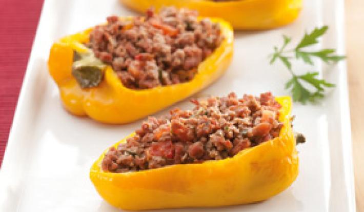Fotografia em tons de vermelho e amarelo de uma bancada de madeira clara, com um pano vermelho dobrado, sobre ele um prato retangular branco com três metades de pimentões amarelos recheados com carne. Sobre o prato branco, 3 folhas de salsa.