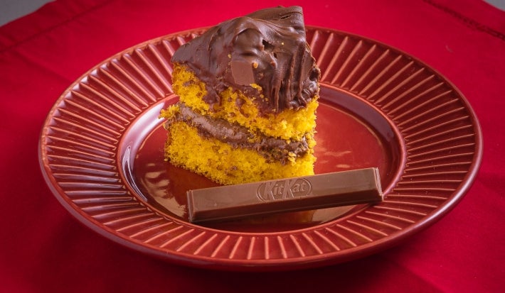 Fotografia de um pedaço de bolo de cenoura com recheio e cobertura de ganache de kiktat em um prato vermelho com um kitkat sobre um pano vermelho.