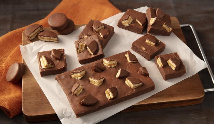 Fotografia em tons de preto e laranja de uma bancada preta com um paninho laranja, sobre ele uma tabua de madeira com um papel manteiga e o fudge de bono doce de leite. Ao lado pedaços do fudge e biscoitos bono coberto espalhados.
