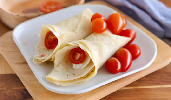 Fotografia em tons de bege e azul com um prato branco ao centro. Dentro do prato existe dois enrolados de crepe recheados com creme de palmito com NINHO Primeira Infância e tomates cereja.