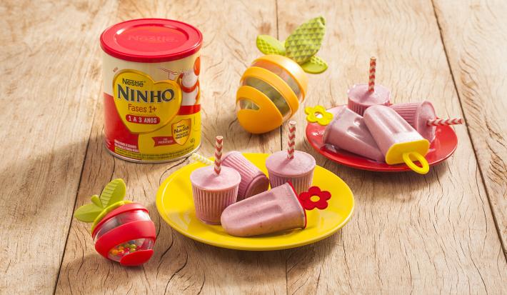 Fotografia em tons de amarelo e vermelho em uma bancada de madeira clara, dois pratos vermelho e amarelo pequenos com picolés pequenos de banana e morango. Ao lado, um lata de Ninho Fases e brinquedinhos infantis.