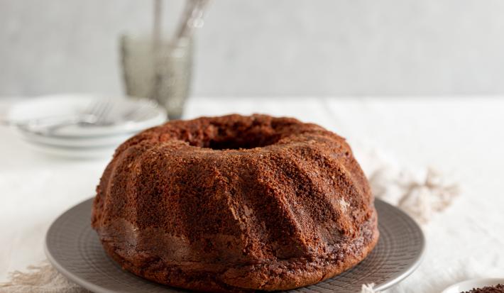 Fotografia de um bolo de chocolate feito em forma com furo central sobre um prato raso de vidro cinza. Ao lado tem granulado de chocolate em um prato pequeno raso. No fundo tem pratos brancos e garfo, e o bolo está sobre um pano branco.