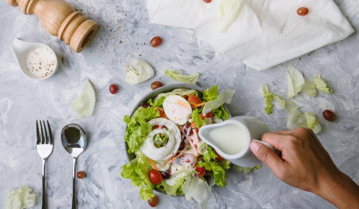 Fotografia vista de cima de uma mão segurando uma jarra pequena na cor branca, que está caindo um molho branco em uma salada de folhas, cebola, ovo cozido, maçã, tomates cereja, cenoura e azeitona preta. O refratário está sobre uma mesa de mármore branca.