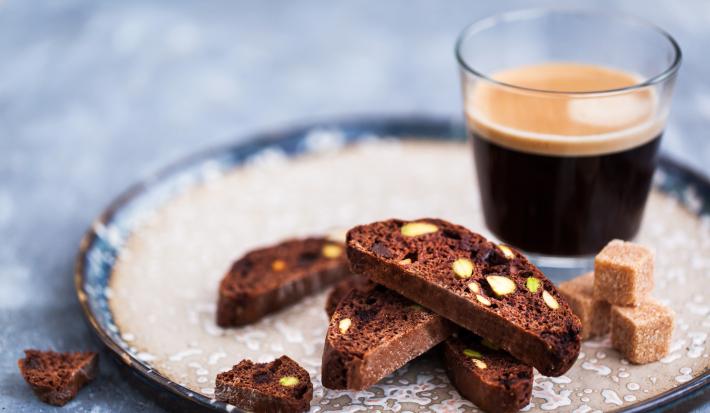 Fotografia quatro fatias de um torrão de chocolate ao lado de cubinhos de açúcar, para acompanhar um Nescafé Espresso Intenso que está em uma xícara de vidro, sobre um prato de vidro desenhado, de cora branca e azul.