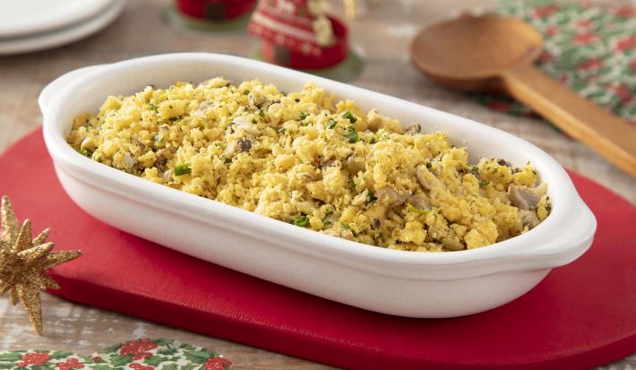 Foto da receita de Farofa Vegana de Legumes. Observa-se um refratário branco com a farofa dentro sobre um guardanapo vermelho. Itens de decoração de Natal decoram a foto.