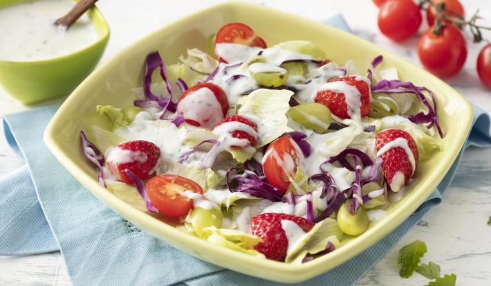 Fotografia em tons de branco, azul e verde de uma bancada branca e um paninho azul, sobre ele um recipiente quadrado verde com a salada e o molho. Ao fundo um recipiente quadrado verde com o molho, uma colher e tomatinhos cereja.