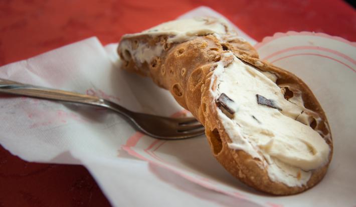 Fotografia de um cannoli de chocolate com creme branco sobre um papel, e ao lado de um garfo.