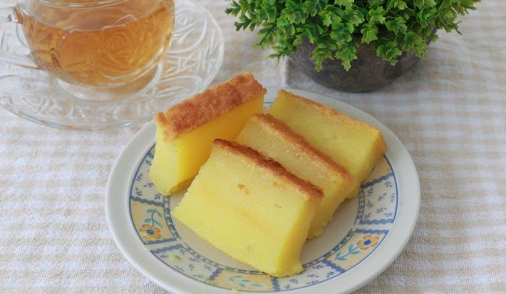 Foto da receita do Bolo de Macaxeira Cremoso. Observa-se um prato de sobremesa com quatro fatias finas do bolo cremoso. Ao lado esquerdo, uma xícara transparente de chá sobre uma toalha quadriculada off white