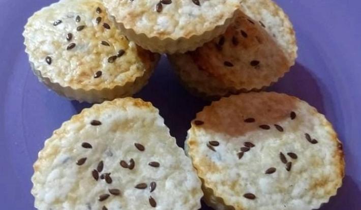 Observa-se 5 pães de queijo feitos em forminhas e polvilhados com semente de linhaça por cima sobre um prato roxo.