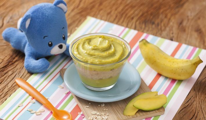 Fotografia em tons de verde em uma bancada de madeira, com um paninho listrado colorido e uma tacinha transparente com abacate e banana apoiado em um pratinho azul. Ao lado, um ursinho de pelúcia azul, uma colher laranja e uma banana.