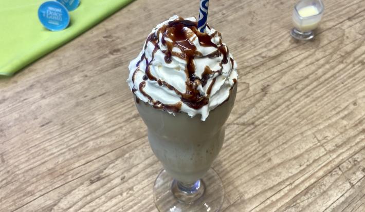 Fotografia em tons de azul e verde em uma bancada de madeira com um copo de vidro alto ao centro e a raspadinha dentro com calda de chocolate e um canudo de papel.