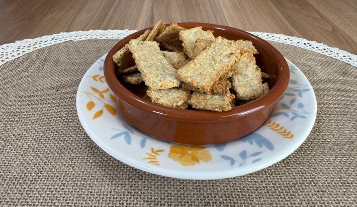 Fotografia de um prato de vidro raso branco com cores claras, sobre ele tem um recipiente pequeno fundo de tom marrom e redondo com vários biscoitinhos de aveia com queijo.