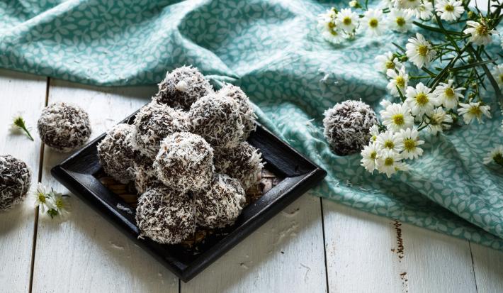 fotografia em tons de marrom, cinza e azul de uma bancada cinza vista de frente. Contém um prato quadrado preto com as balas de chocolate cobertas por coco ralado, e ao lado um pano azul com flores brancas.
