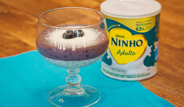 Fotografia em tons de azul com uma taça de vidro ao centro. Dentro da taça existe um doce em camadas feito com mirtilos, creme de ninho e chias. Ao lado existe um lata de leite NINHO Adulto