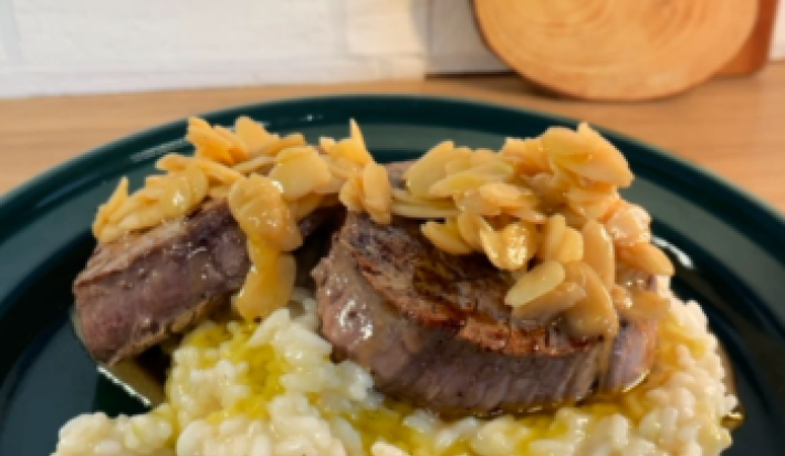 imagem mostra um prato de risoto cremoso em tons beges e por cima 2 medalhões de filé mignon com amendoas laminadas e um molho amanteigado por cima.