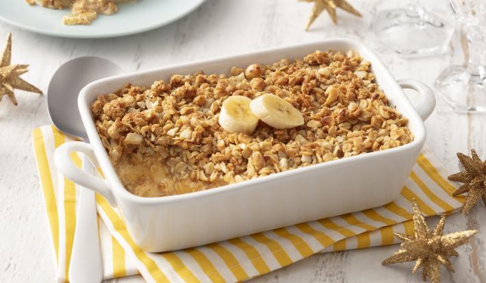 Fotografia em tons de branco e amarelo de uma bancada branca com um paninho listrado amarelo e branco, sobre ele um recipiente retangular branco com o crumble e uma colher. Ao fundo um prato com um pedaço de crumble.