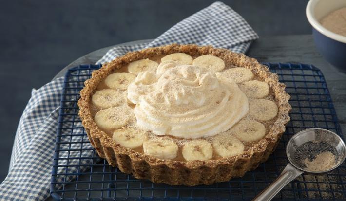 Foto da receita de banoffee de farinha lactea aveia servida em uma porção grande sobre uma grade preta com uma colher de servir prateada ao lado