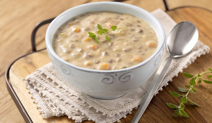 Foto em tons de amarelo com a receita de creme de lentilha, requeijão NESTLÉ e peito de peru servida em um bowl branco com adornos sobre um pano de renda bege em cima de uma tábua de madeira e uma colher prateada à frente. Tudo em cima da mesa de madeira.