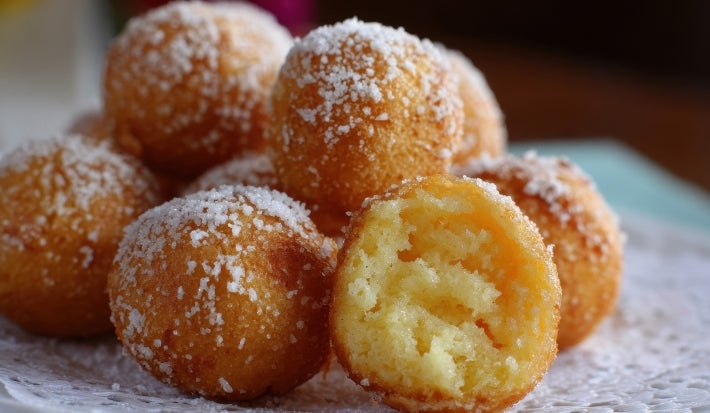 Fotografia de um prato com bolinhas de massa frita, cobertas de açúcar, com uma aparência dourada e crocante. Uma delas está cortada, mostrando um interior macio e fofo, dispostas sobre um delicado guardanapo de renda.