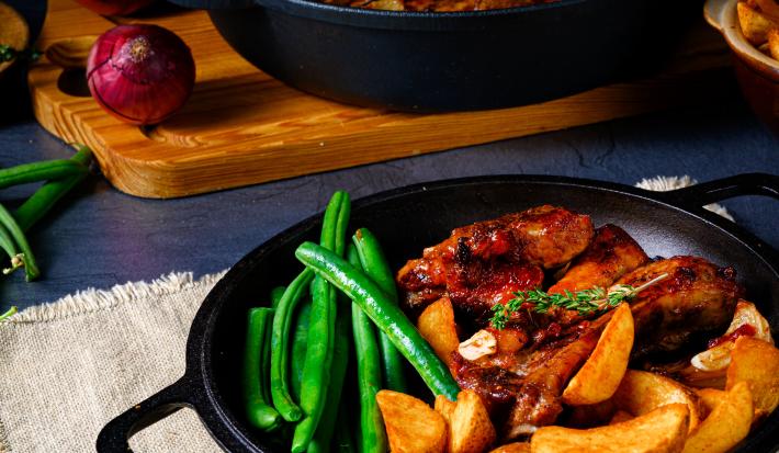 Fotografia de alguns pedaços de frango assado com batatas, vagens e ervilha torta dentro de uma panela rasa de ferro na cor preta, que está em cima de um pano branco. Ao fundo, outra panela de ferro preta com o resto dos frangos assados sobre uma tábua.