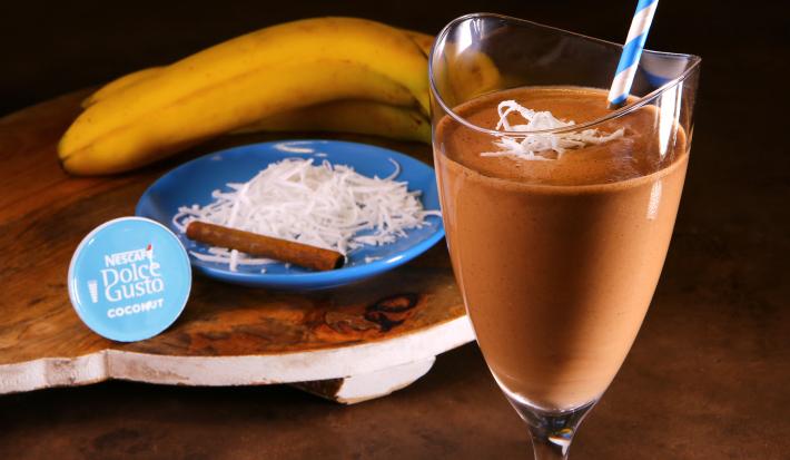 Fotografia em tons de azul em uma bancada de madeira, uma tábua de madeira com um copo de vidro alto e a bebida de chocolate com coco e banana dentro dele. Ao fundo, um prato azul com raspas de coco, uma canela em pau e uma banana.