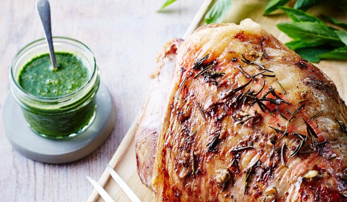 Fotografia de um pernil de cordeiro temperado com laranja e molho de hortelã ao lado em um pote de vidro, com uma mini colher apoiada. O pernil está sobre uma tábua de madeira, ao lado de algumas folhas verdes.