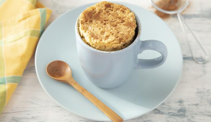 Fotografia em tons de azul em uma bancada de madeira clara, um prato e uma xícara azul ao centro com o bolo de caneca dentro. Ao lado, um pano amarelo e uma peneirinha com canela.