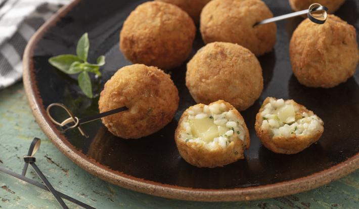 Imagem de um prato rústico preto com diversos bolinhos arancini e um bolinho ao meio mais a frente.