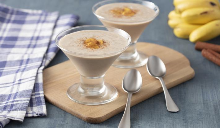 Fotografia em tons de rosa de uma bancada cinza, ao centro uma tábua de madeira com duas taças com um creme bege decorado com banana. Ao lado duas colheres prateadas.