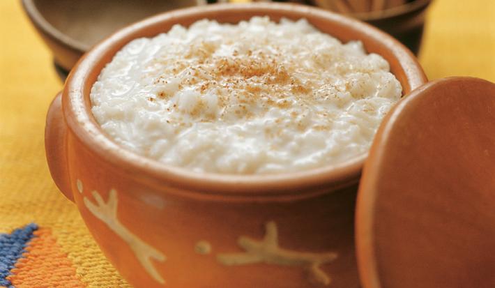 Fotografia em tons de amarelo em uma mesa de madeira com uma toalha amarela desenhada, um recipiente de barro com o arroz doce dentro dele. Ao fundo, um potinho com paus de canela.