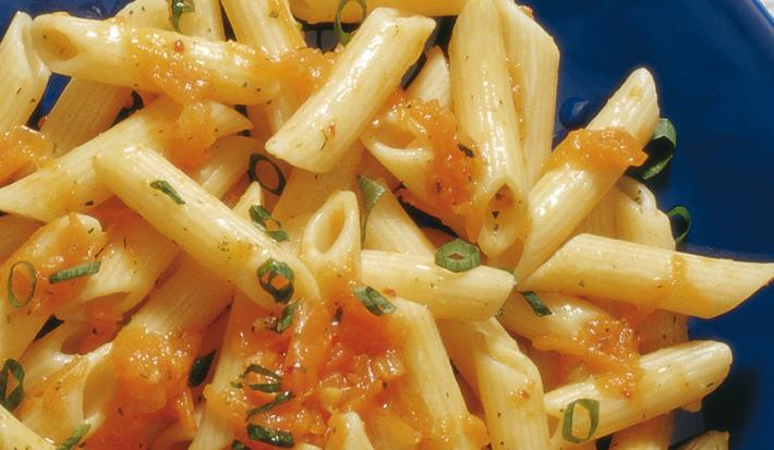 Fotografia em tons de amarelo e azul, com destaque para porção de macarrão cozido com pedaços de abóbora e cebolinha dentro de um prato azul, sobre toalha em tom de azul.