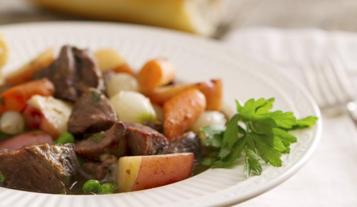 Fotografia em tons de branco, marrom, amarelo e laranja de uma bancada branca vista de frente. Contém um prato redondo branco, e dentro picadinho de carne com batatas e cenouras em cubos.
