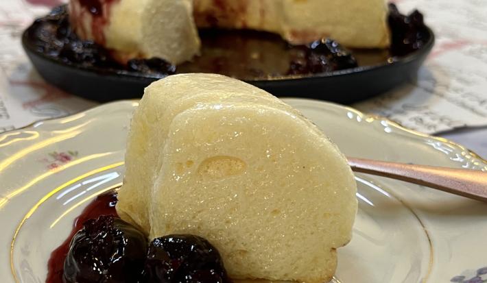Foto da Receita de Pudim de Claras. Observa-se um prato de sobremesa branco de porcelana com uma fatia do pudim e calda de ameixas. Atrás, o pudim inteiro com a calda.