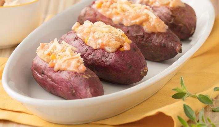 Fotografia em tons de amarelo em uma bancada de madeira com um pano amarelo, um recipiente oval branco com as batatas-doces recheadas com frango cremoso.