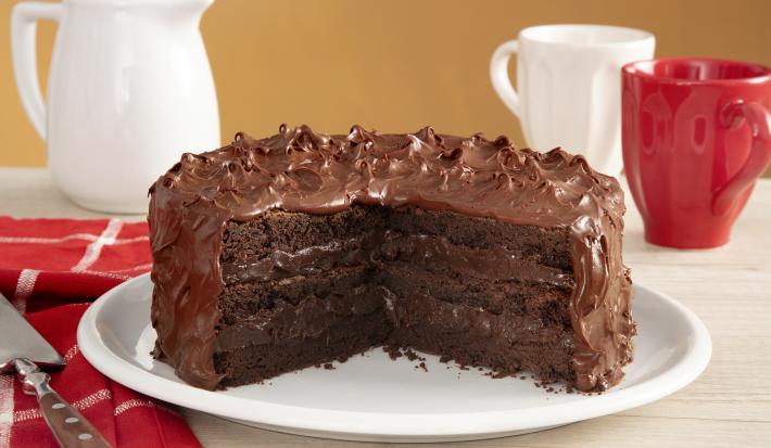 Fotografia em tons de vermelho em uma bancada de madeira com um pano vermelho listrado em branco, com um prato branco no centro com o bolo brownie com brigadeiro de cacau de cobertura. Ao fundo, uma xícara branca, uma xícara vermelha e um bule branco.