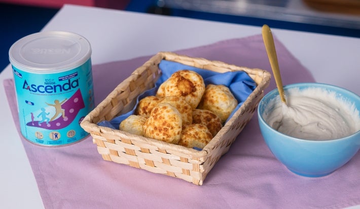 Fotografia em tons claros de uma cestinha de madeira retangular sobre um pano quadrado roxo. Dentro da cestinha tem um paninho menor azul com os pães de queijo, ao lado tem uma lata de Ascenda e um recipiente azul fundo pequeno com o creme de inhame.