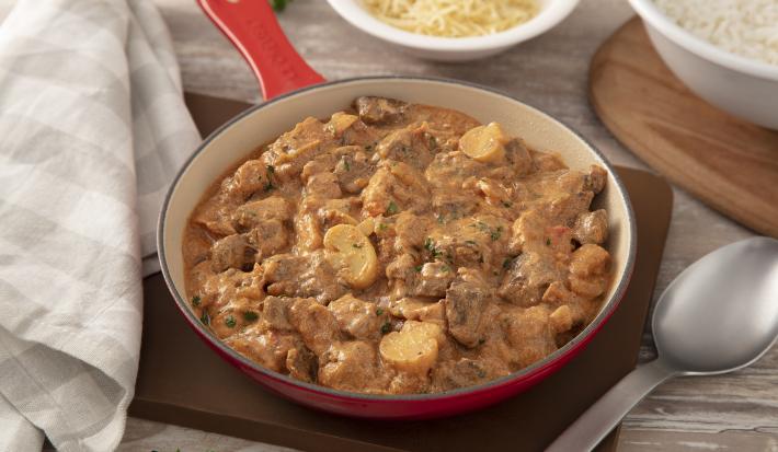 Foto da receita de strogonoff de carne. Observa-se uma panela grande com o strogonoff sobre uma tábua de madeira. Ao lado, um colher de servir e duas tigelas: uma com batata-palha e outra com arroz.