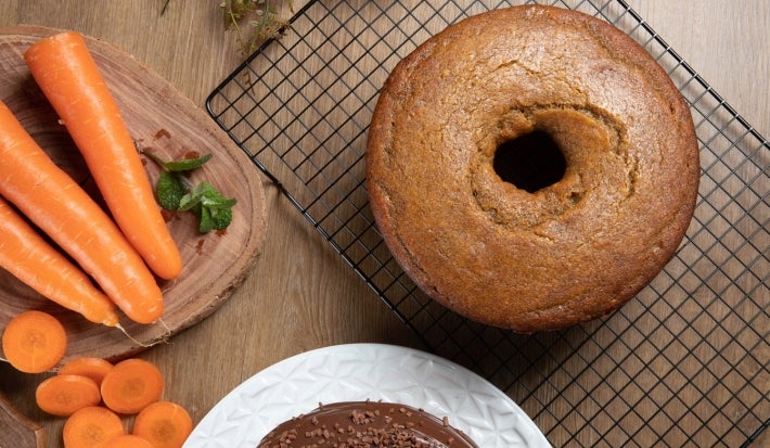Bolo de cenoura com furo no centro, assado e dourado, resfriando sobre uma grade. Ao lado, cenouras frescas e um bolo decorado com cobertura de chocolate e granulados.
