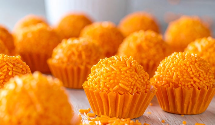 Fotografia mostra brigadeiros laranjas em forminhas laranja sobre uma mesa de madeira.