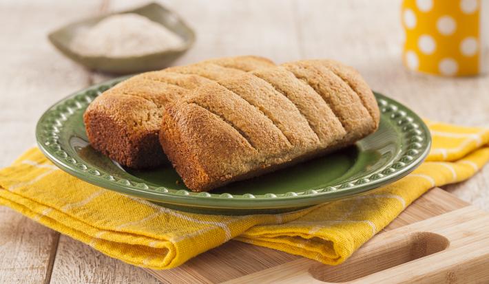 Fotografia em tons de amarelo em uma bancada de madeira clara, um pano amarelo com listras brancas, uma tábua de madeira, um prato verde redondo com dois pãezinhos em cima dele. Ao fundo, uma xícara amarela com bolinhas brancas.