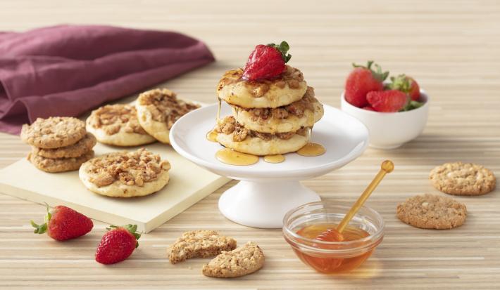 Foto de uma bancada de madeira clara. Sobre ela há morangos soltos e em um pote, cookies em uma tábua, um pote de vidro com mel e um guardanapo vinho. Ao centro há um prato com três pancakes cobertas com mel e decoradas com morangos.