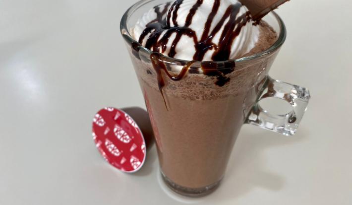 Fotografia em tons de branco com uma caneca de vidro ao centro. Dentro da caneca existe uma receita de frappuccino de cor chocolate coberto com chantilly e calda de chocolate. Ao lado existe uma cápsula de NESTLÉ Dolce Gusto KitKat