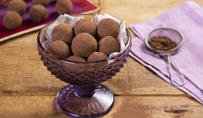 fotografia em tons de marrom e lilás de uma bancada marrom vista de cima, contém uma taça lilás com trufas redondas de chocolate e ao lado um pano lilás com uma peneira que contém cacau em pó.