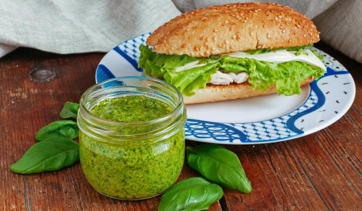 Fotografia de uma mesa de madeira e, sobre ela, um pote de vidro pequeno de vidro com pesto dentro. Ao lado tem u prato branco com detalhes azuis, apoiado no prato tem um sanduíche de frango com um pouco do pesto de coentro.