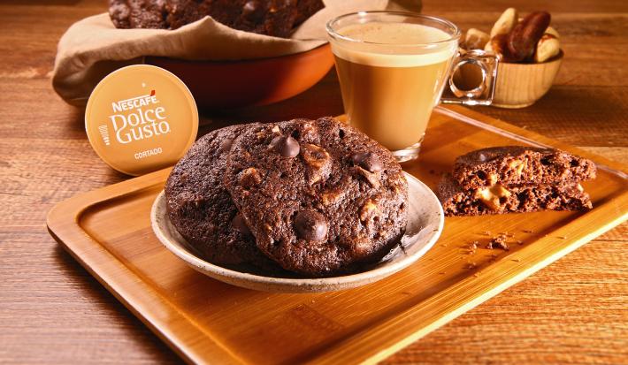 Fotografia em tons de bege em uma bancada de madeira escura, uma tábua de madeira com um pratinho bege redondo e alguns cookies de chocolate com café em cima dele. Ao lado, uma xícara de vidro com o Café Cortado Dolce Gusto e ao fundo mais cookies.