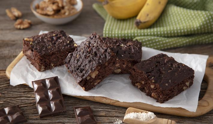 Fotografia em tons de marrom em uma bancada de madeira de cor marrom. Ao centro, uma tábua de madeira contendo os brownies em cima. Ao lado, há alguns tabletes de chocolate e ao fundo, um pano verde quadriculado com algumas bananas em cima.