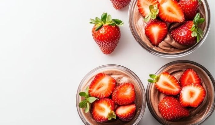 Fotografia em tons claros, vista de cima, de três recipientes de vidro pequenos com a sobremeza dentro, finalizados com ganache e morangos cortados pela metade. Os potinhos estão sobre uma mesa branca, ao lado dos potinhos tem morangos inteiros.