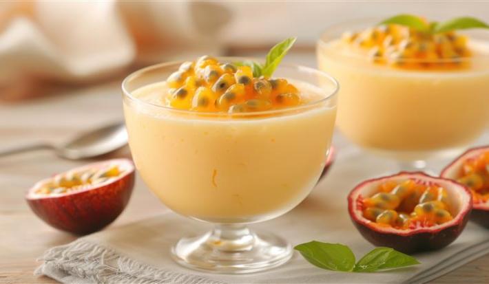 Fotografia de dois recipientes pequenos de vidro com mousse de maracujá e a polpa por cima. Os potinhos estão sobre uma mesa clara, com várias metades de maracujá.