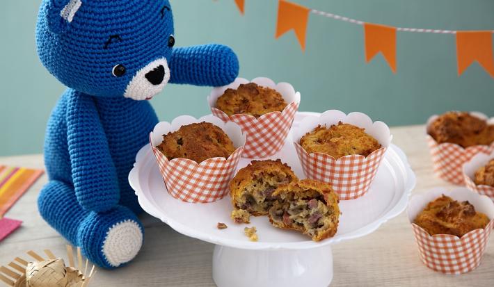 Fotografia em tons de laranja, em uma bancada de madeira de cor branca. Ao centro, uma boleira contendo os muffins com um ursinho azul, um mini chapeu de palha e um pano listrado colorido ao lado. Ao fundo, uma parede azul piscina e mini bandeiras.